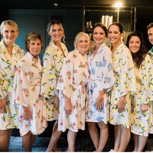 May include: A group of women wearing white robes with floral patterns. The robes have different floral designs, including lemons, flowers, and hydrangeas. The women are smiling and looking at the camera.