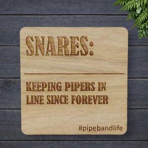 May include: A square wooden sign with rounded corners. The sign has the words "SNARES: KEEPING PIPERS IN LINE SINCE FOREVER" engraved in a dark brown font. The hashtag "#pipebandlife" is at the bottom.