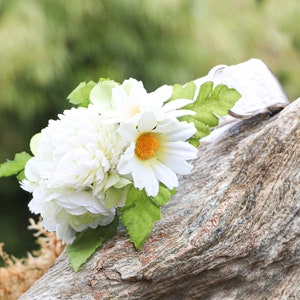 May include: White flower collar with white daisies and green leaves. The collar is made of a white fabric and is decorated with white daisies and green leaves. The collar is lying on a piece of wood.