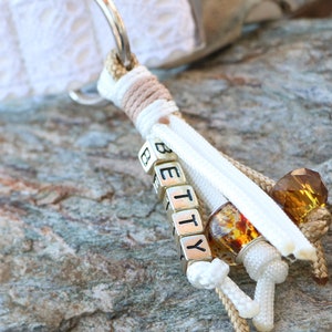 May include: White and beige rope keychain with a silver charm that spells "BETTY" in block letters. The charm is attached to a white and beige rope with amber-colored beads.