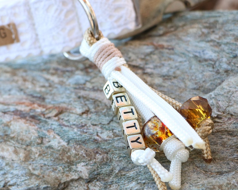 May include: White and beige rope keychain with a silver charm that spells "BETTY" in block letters. The charm is attached to a white and beige rope with amber-colored beads.