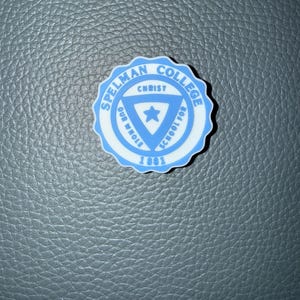 May include: A round, light blue and white Spelman College emblem. The emblem features the text "Spelman College" and "Christ Our Whole School For 1881" in a circular design. A star is in the center.
