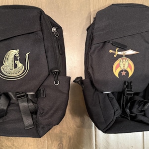May include: Two black backpacks with different designs. The backpack on the left has a gold design of an Egyptian woman with a crown. The backpack on the right has a gold design of a crescent moon, star, and sword.