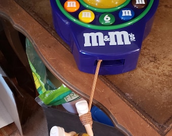 Könnte beinhalten: Ein blau-grünes M&M's-Spielzeugtelefon mit einer gelben M&M's-Figur, die an einer Schnur hängt. Das Telefon hat ein Ziffernblatt mit Zahlen und das M&M's-Logo.