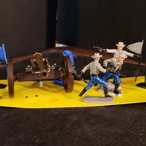 May include: A toy Civil War battle scene with miniature soldiers, a cannon, flags, and a horse. The figures are in blue and gray uniforms, with a brown barrel and a yellow base. One flag has stars and stripes.