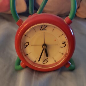 May include: A round analog alarm clock with a bright red frame and a white face featuring black numerals at the 12, 3, 6, and 9 positions. The clock has thin black hands and is supported by curved teal green legs that extend upward from the base.