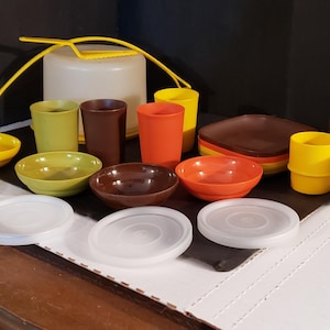 May include: A collection of vintage plastic dishware in various colors, including yellow, orange, brown, and green. The set includes bowls, cups, mugs, and lids. A white container with a yellow cord is also present.