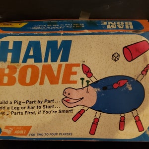 May include: A vintage board game box with a blue and orange design. The box features the game's name, "Ham Bone", and an illustration of a pig with a blue body and pink snout. The game is for two to four players and is recommended for ages 4 and up.