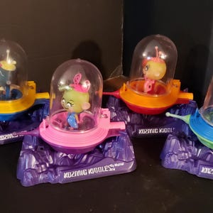 May include: Four Kosmic Kiddles toys by Mattel, each featuring a small doll inside a clear plastic dome on a colorful spaceship base. The bases are yellow, pink, orange, and blue, all mounted on purple rocky terrain stands with printed text.