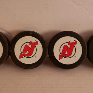 May include: Four black hockey pucks with a white center and a red New Jersey Devils logo.