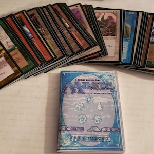 May include: A collection of Magic: The Gathering trading cards, featuring various card designs and colors. A deck box with the text "Magic: The Gathering Ice Age" is also present. The cards are arranged in stacks.