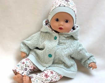 May include: A doll wearing a light mint green hooded jacket with a cable knit texture and two teal buttons, paired with matching floral print leggings and a coordinating floral beanie hat. The doll has a light skin tone and dark brown eyes.