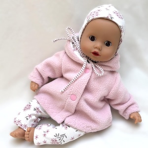 Könnte beinhalten: Eine Puppe trägt eine hellrosa Fleecejacke mit Kapuze und zwei rosa Knöpfen, eine weiße Hose mit kleinem rosa-grauem Blumenmuster sowie eine passende weiße Mütze mit rosa-grauem Blumenmuster und gestreiften Bändern unter dem Kinn.