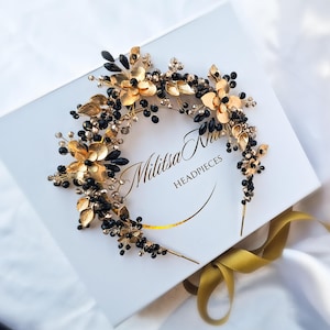 Könnte beinhalten: Ein gold-schwarzes Blumen-Haarband mit dem Text "Militsa Knai HEADPIECES" auf einer weißen Schachtel. Das Haarband hat goldene Blumen, Blätter und schwarze Perlen. Ein goldenes Band ist sichtbar.