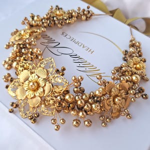 Könnte beinhalten: Ein goldener Braut-Haarschmuck mit floralem Design. Der Haarschmuck besteht aus goldenen Perlen, Blumen und Draht. Der Haarschmuck befindet sich auf einem weißen Hintergrund mit dem Text "Headpieces" und "Daisy Lane".