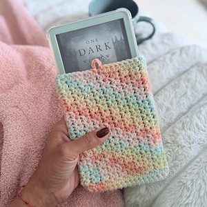 May include: A pastel-colored, crocheted Kindle sleeve holds an e-reader displaying the book cover of "One Dark Window." The sleeve is made of yarn in shades of pink, yellow, green, and blue. A small loop is attached at the top. A mug is in the background.