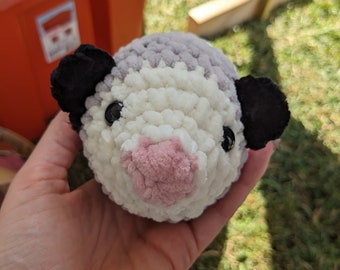 May include: A crocheted stuffed animal resembling a possum with a white and gray body, black ears, and a pink nose.