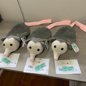 K&ouml;nnte beinhalten: Drei graue und wei&szlig;e geh&auml;kelte Opossum-Pl&uuml;schtiere mit rosa Schw&auml;nzen. Jedes Pl&uuml;schtier hat schwarze Augen und eine rosa Nase. Die Pl&uuml;schtiere sitzen auf einem Holztisch mit wei&szlig;en Karten, auf denen "Crafty Chick Crochet" und "Follow" steht.