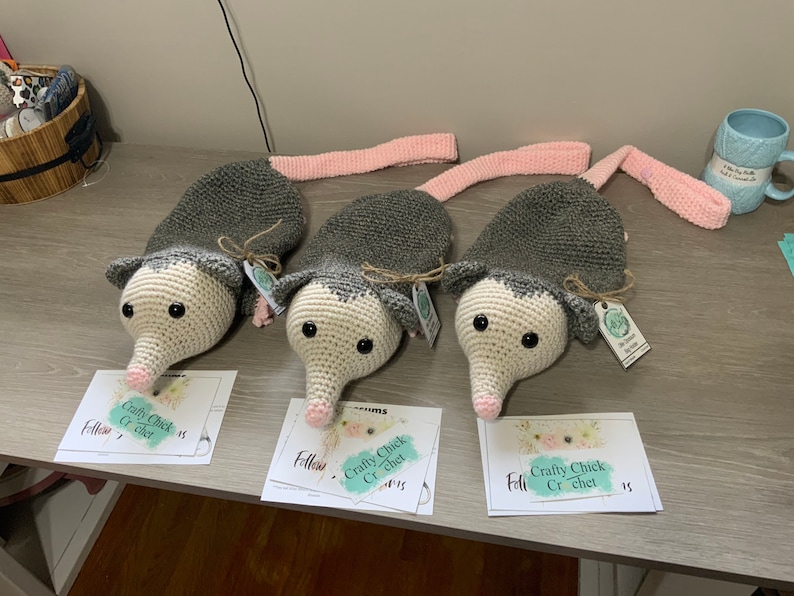 K&ouml;nnte beinhalten: Drei graue und wei&szlig;e geh&auml;kelte Opossum-Pl&uuml;schtiere mit rosa Schw&auml;nzen. Jedes Pl&uuml;schtier hat schwarze Augen und eine rosa Nase. Die Pl&uuml;schtiere sitzen auf einem Holztisch mit wei&szlig;en Karten, auf denen "Crafty Chick Crochet" und "Follow" steht.