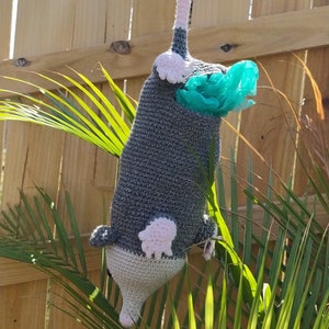K&ouml;nnte beinhalten: Ein graues, geh&auml;keltes Stofftier in Form eines Opossums mit rosa Ohren und einem Schwanz. Das Opossum h&auml;lt einen blauen Plastikbeutel im Maul und h&auml;ngt an einem rosa geh&auml;kelten Band.