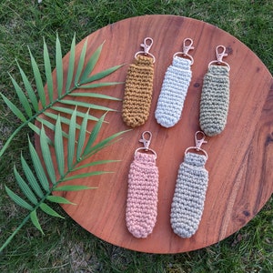 May include: Six crocheted lip balm holders with rose gold keychains. The holders are in various colors, including mustard yellow, white, sage green, light pink, and gray.