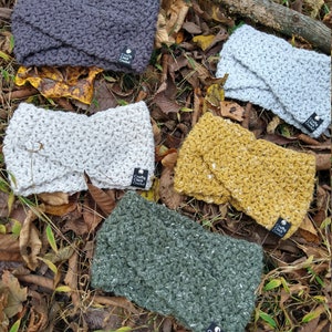 May include: Five crocheted headbands in different colors and textures, including gray, white, yellow, green, and a light gray. Each headband has a small black tag with the text "Crafty Chick Crochet" on it.