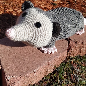 K&ouml;nnte beinhalten: Ein geh&auml;keltes graues und rosa Opossum-Pl&uuml;schtier mit schwarzen Augen. Das Spielzeug sitzt auf einem Satz brauner Ziegel.