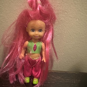 May include: A doll with pink hair wearing a green and pink outfit with a jeweled necklace. The doll has brown eyes and a painted smile.