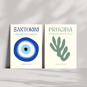 May include: Two art prints on a cream background. The left print says "SANTORINI" above "AEGEAN SEA, GREECE" and features a blue eye design. The right print says "PROCIDA" above "TYRRHENIAN SEA, ITALY" and has a green abstract shape. Both prints say "BEACH CLUB".