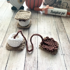 May include: A crocheted s'mores set featuring a brown marshmallow, graham cracker, and chocolate. The set includes a white marshmallow, a brown chocolate square, and a graham cracker square. The s'mores are connected by a brown crocheted rope.