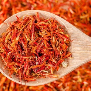 May include: A wooden spoon filled with dried safflower petals, a vibrant orange-red colour. The petals are arranged in a loose pile, with some visible stems.