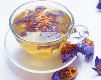 May include: A glass cup with a clear tea infuser filled with a purple flower tea. The tea is a light yellow color. There are several purple flower petals scattered around the cup.