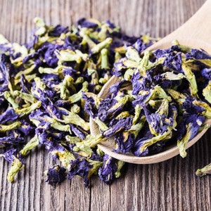 May include: A wooden spoon filled with dried blue butterfly pea flowers, with more flowers scattered on a wooden surface.