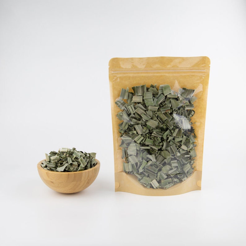 May include: A brown paper pouch with a clear plastic window filled with dried green lemongrass. A small wooden bowl filled with dried lemongrass sits in front of the pouch.