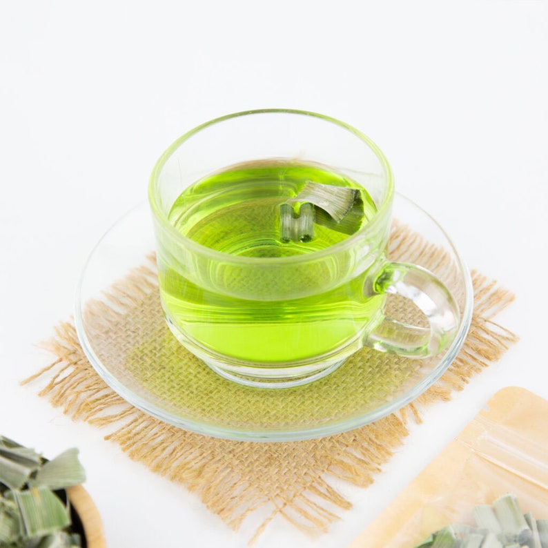 May include: A clear glass cup filled with green lemongrass tea on a brown woven placemat. The tea has a few pieces of lemongrass floating in it.