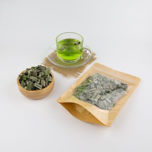 May include: A brown paper bag with a clear window showing dried green lemongrass inside. A wooden bowl filled with dried lemongrass sits next to the bag. A glass cup of green lemongrass tea sits on a brown woven coaster.
