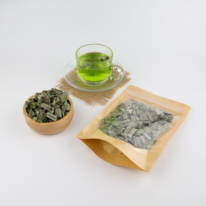 May include: A brown paper bag with a clear window showing dried green lemongrass inside. A wooden bowl filled with dried lemongrass sits next to the bag. A glass cup of green lemongrass tea sits on a brown woven coaster.
