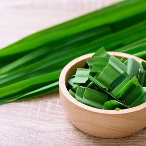 May include: A wooden bowl filled with chopped green pandan leaves. The leaves are a vibrant green and are arranged neatly in the bowl. The bowl is sitting on a light brown wooden surface.