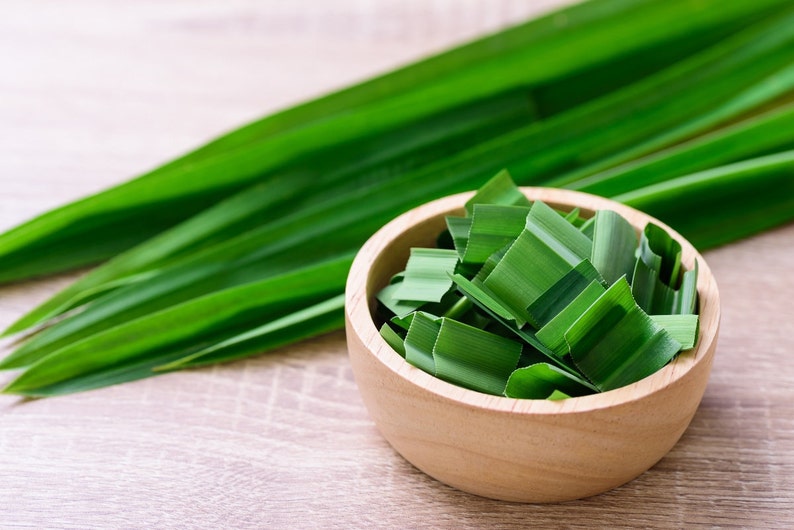 May include: A wooden bowl filled with chopped green pandan leaves. The leaves are a vibrant green and are arranged neatly in the bowl. The bowl is sitting on a light brown wooden surface.