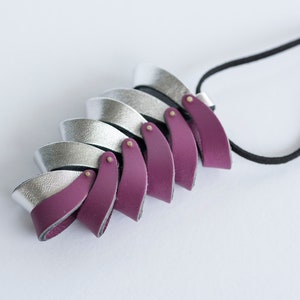 Könnte beinhalten: Eine silberne und burgunderfarbene Lederkette mit geometrischem Design. Die Kette besteht aus mehreren übereinanderliegenden Lederstücken, die jeweils eine andere Farbe und Textur haben. Die Kette ist an einer schwarzen Schnur befestigt.