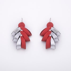 Puede incluir: Un par de pendientes hechos a mano con una parte superior circular roja y un diseño en forma de abanico de piezas de cuero plateadas y rojas alternas. Los pendientes tienen una estética moderna y geométrica y están suspendidos de ganchos plateados.