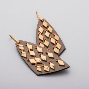 May include: A pair of dangle earrings. Each earring features a gray, leaf-shaped base adorned with gold diamond-shaped accents and small orange beads. The earrings have gold-colored ear wires.