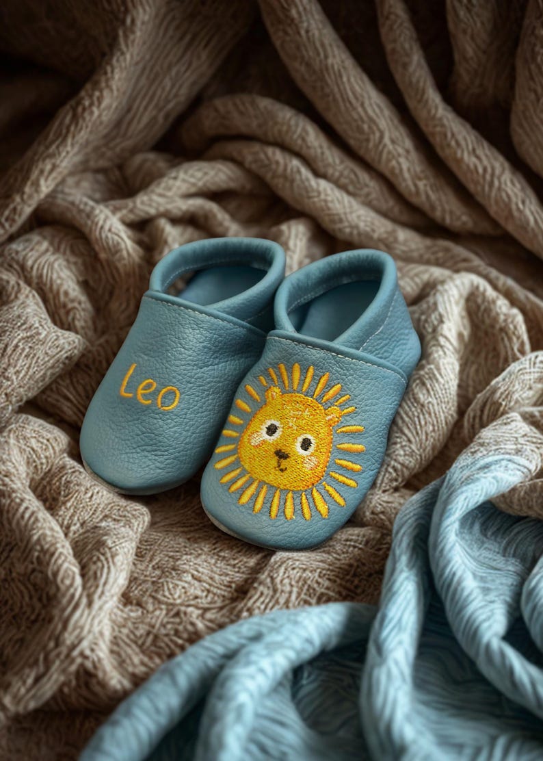 May include: A pair of light blue soft-soled baby shoes. One shoe features the embroidered name Leo in yellow thread, while the other shoe displays a yellow and orange embroidered lion face with a sun-like mane on the front toe area.