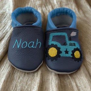 Könnte beinhalten: Ein Paar dunkelblaue Leder-Babyschuhe mit hellblauem Rand. Ein Schuh ist mit dem Namen Noah in hellblauem Garn bestickt, während der andere Schuh einen gestickten hellblauen Traktor mit gelben Rädern und einer kleinen weißen Rauchwolke zeigt.
