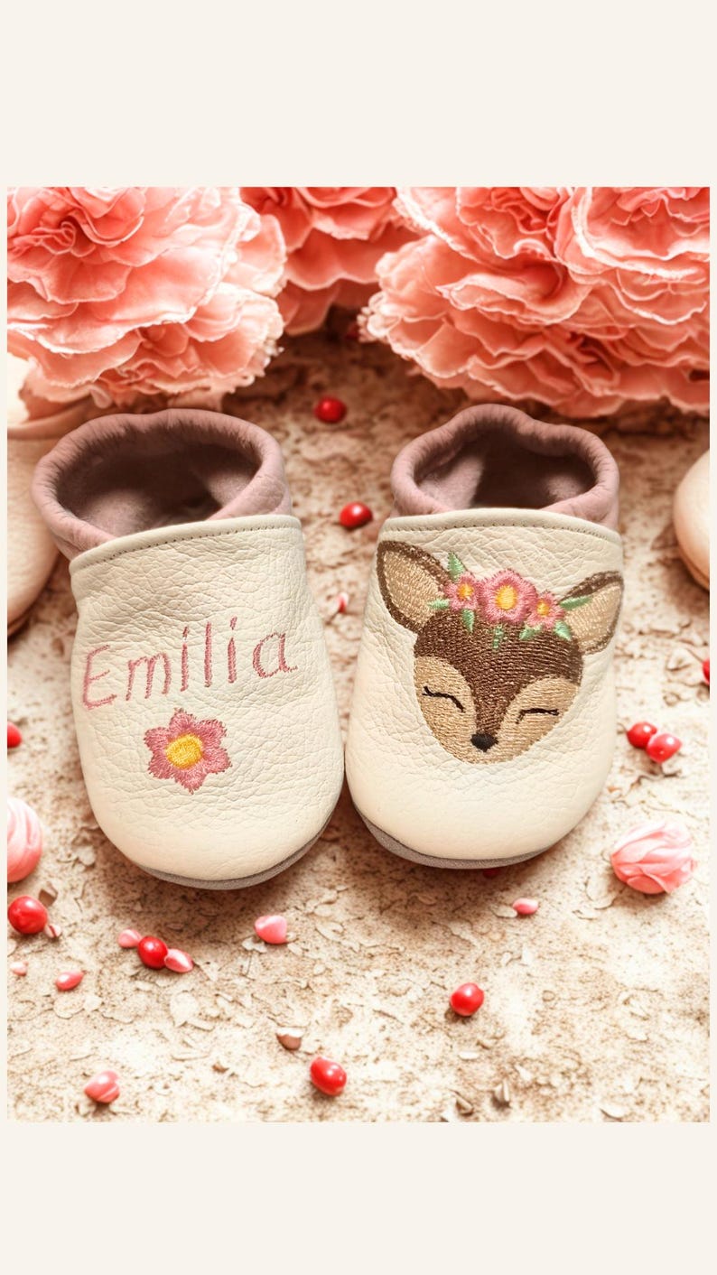 May include: A pair of cream-colored leather baby slippers with a soft mauve interior. The left slipper features the embroidered name Emilia and a pink flower. The right slipper displays an embroidered deer face with a floral crown.