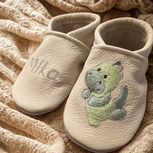 Könnte beinhalten: Ein Paar cremefarbene Leder-Babyschuhe. Ein Schuh ist mit dem Namen Mika in dunklem Garn bestickt, während der andere auf der Vorderseite eine grüne und hellgrüne Stickerei eines Dinosauriers aufweist.
