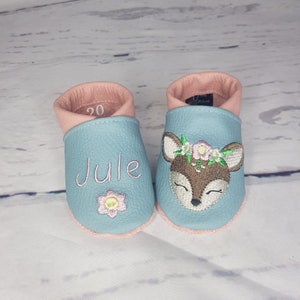 May include: A pair of light blue leather baby shoes with pink trim. One shoe features the embroidered name JULE above a small flower. The other shoe displays an embroidered deer head with a floral crown. The number 20 is visible inside one shoe.