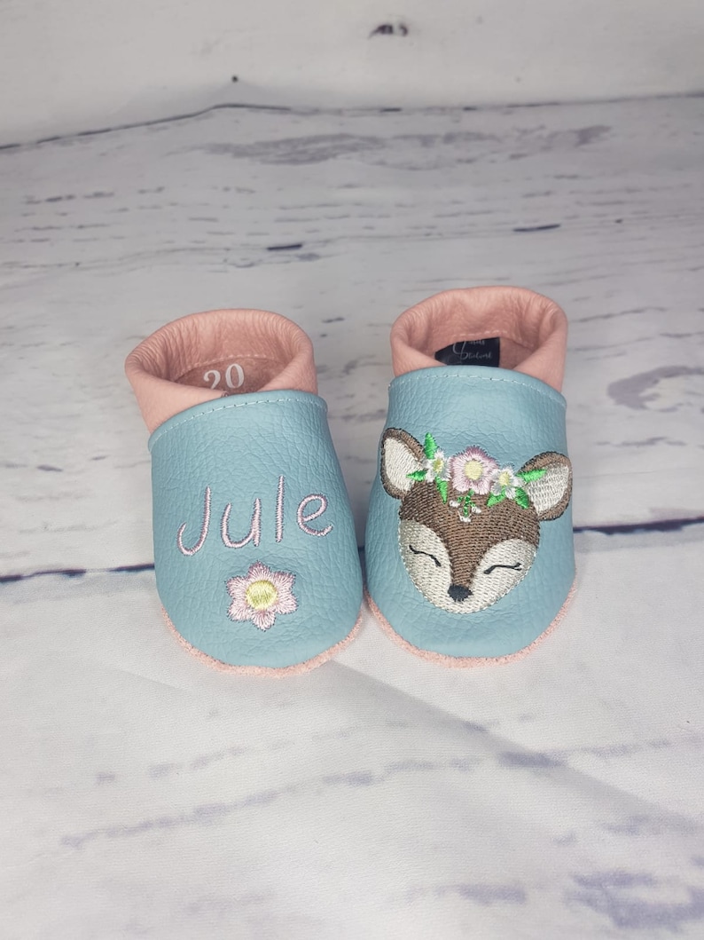 May include: A pair of light blue leather baby shoes with pink trim. One shoe features the embroidered name JULE above a small flower. The other shoe displays an embroidered deer head with a floral crown. The number 20 is visible inside one shoe.