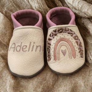 Pode incluir: Um par de sapatos de bebé em pele cor de creme com rebordo rosa. O sapato esquerdo apresenta o nome bordado Adelin, enquanto o sapato direito exibe um desenho de arco-íris bordado com um pequeno coração e detalhes de leopardo.