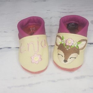 May include: A pair of cream and magenta leather baby shoes. One shoe features the embroidered name Enja in pink with a small flower, while the other displays an embroidered deer face with a floral crown. The interior of the left shoe shows size 18.